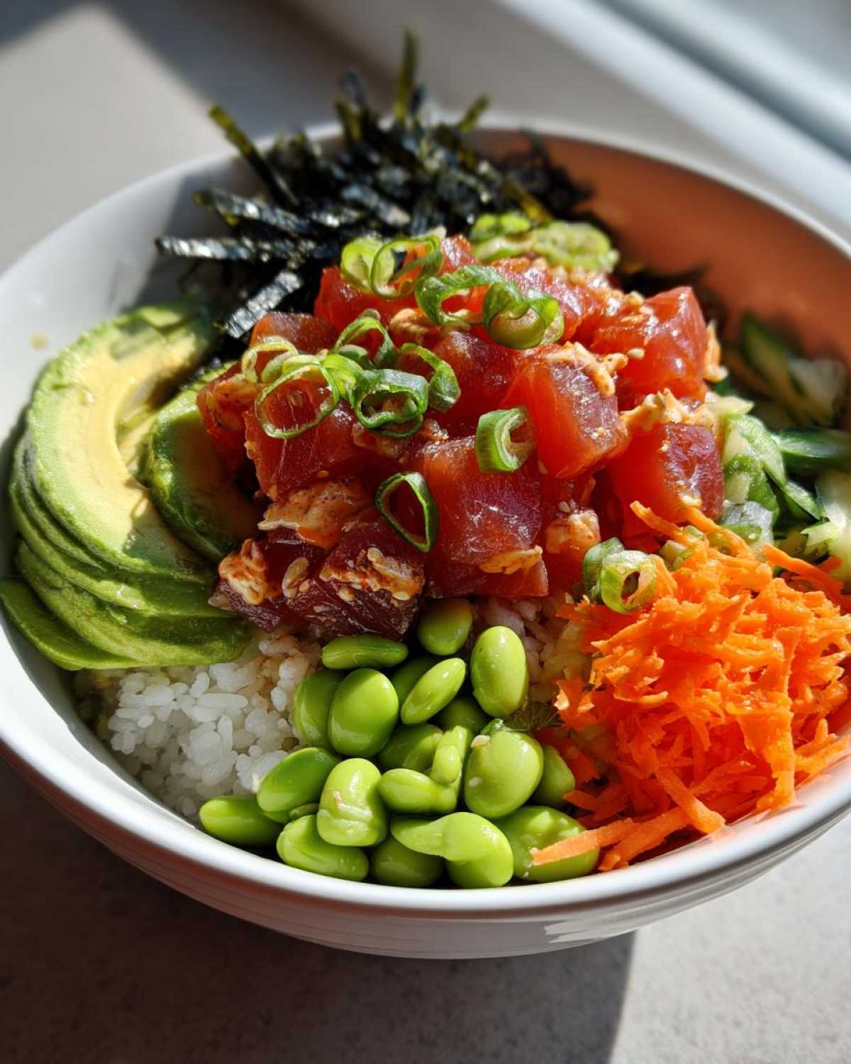A close-up of a homemade Spicy Tuna Poke Bowl featuring marinated tuna, avocado, edamame, shredded carrots, and nori.