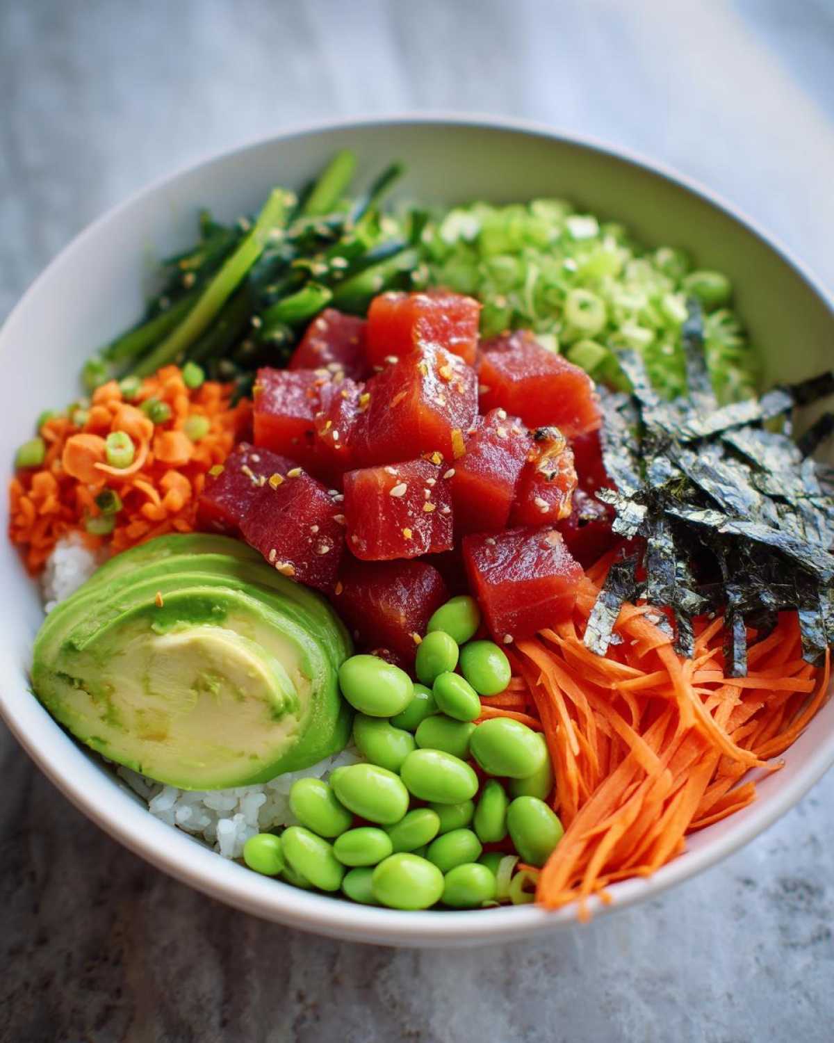 Close-up of a bowl filled with rice, topped with marinated tuna cubes, avocado slices, edamame, carrots, and seaweed for Spicy Tuna Poke Bowls.