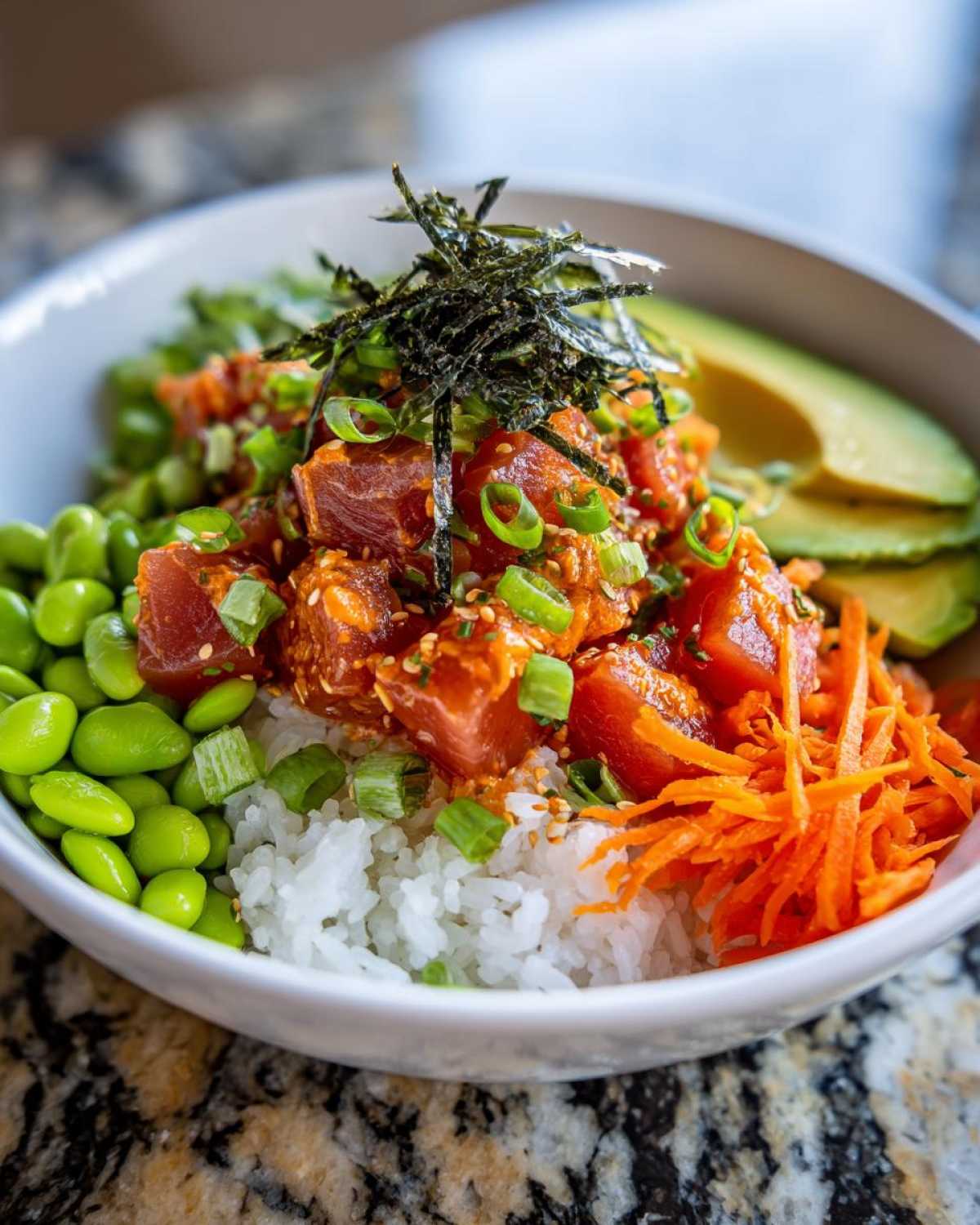 A vibrant bowl featuring Spicy Tuna Poke Bowls ingredients: marinated tuna, white rice, edamame, shredded carrots, avocado slices, and nori.