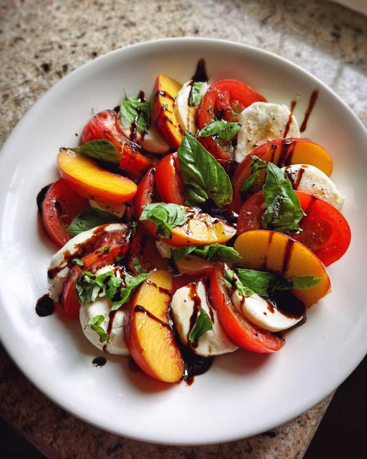 Close-up of a Stone Fruit Caprese Salad featuring sliced peaches, tomatoes, mozzarella, and fresh basil drizzled with balsamic glaze.