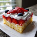 A moist slice of Strawberry Jello Poke Cake Flag topped with whipped cream, fresh strawberries, and blueberries.