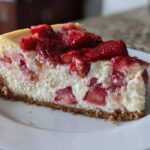 A close-up slice of Strawberry Rhubarb Cottage Cheese Cake showing a thick graham cracker crust and creamy filling studded with strawberries.