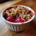 Close-up of a warm, bubbling Strawberry Rhubarb Crisp served in a white ramekin on a wooden surface.