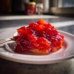 Close-up of a spoonful of vibrant red Strawberry Rhubarb Pineapple Jam resting on a small white plate.