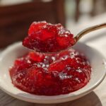 A spoonful of glistening, bright red Strawberry Rhubarb Pineapple Jam being lifted from a small white bowl.