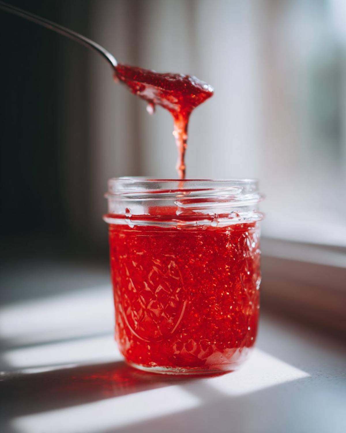 A spoonful of thick, vibrant red Strawberry Rhubarb Soda Syrup drips back into a small glass jar.