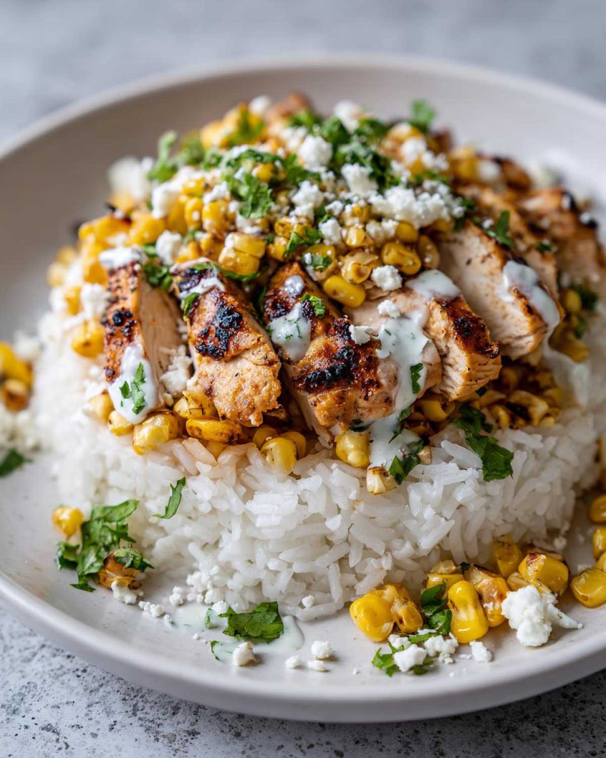 A close-up of a Street Corn Chicken Bowl Recipe featuring sliced grilled chicken over white rice, topped with charred corn, cotija cheese, and cilantro.