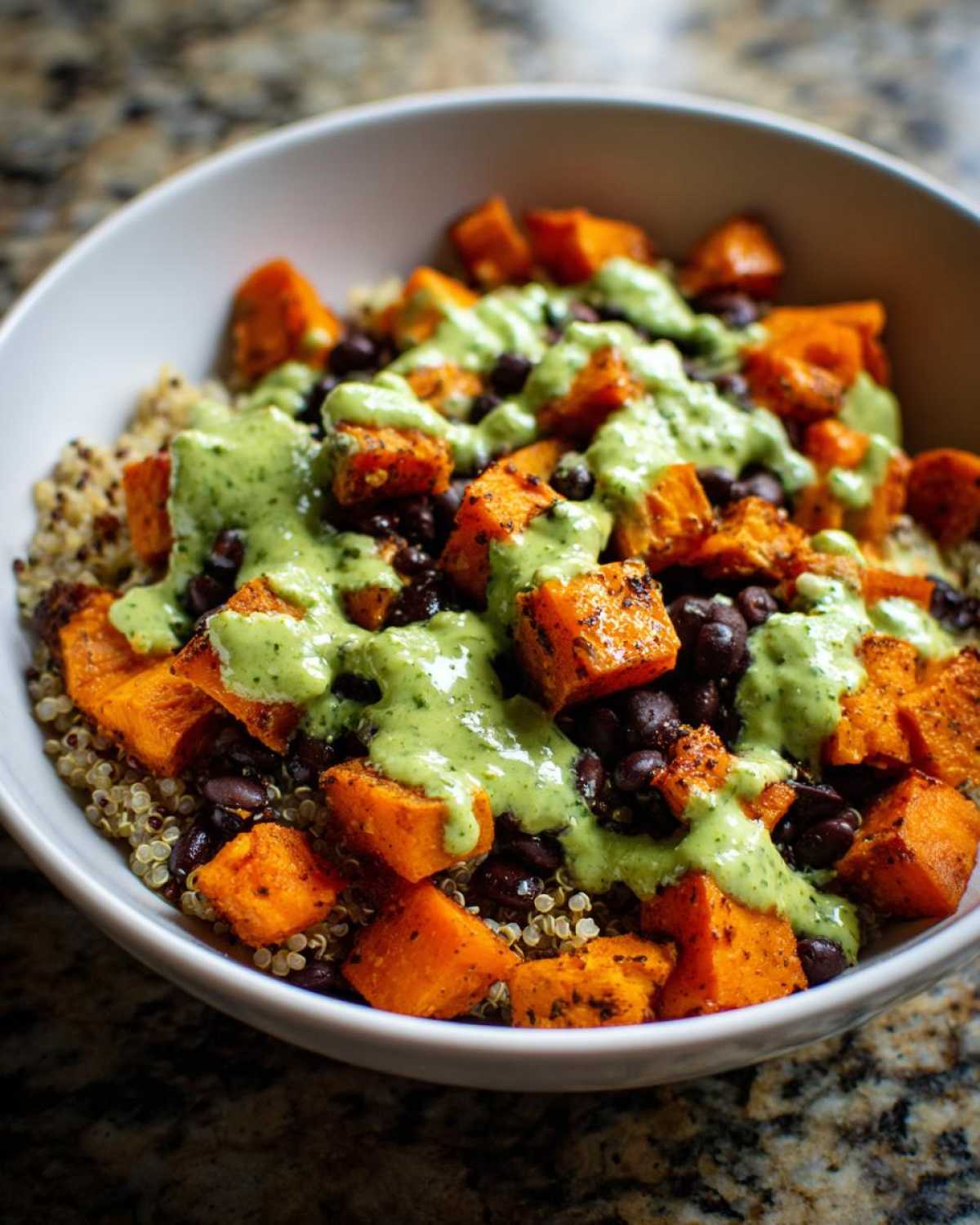 A close-up of a white bowl filled with Sweet Potato & Black Bean Quinoa Bowls, topped with a creamy green sauce.