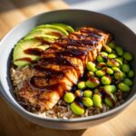 A close-up of a Teriyaki Salmon Bowls featuring glazed salmon, bright green edamame, sliced avocado, and brown rice in a grey bowl.