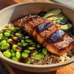 A close-up of a Teriyaki Salmon Bowl featuring a glazed salmon fillet over brown rice, bright green edamame, and sliced avocado.