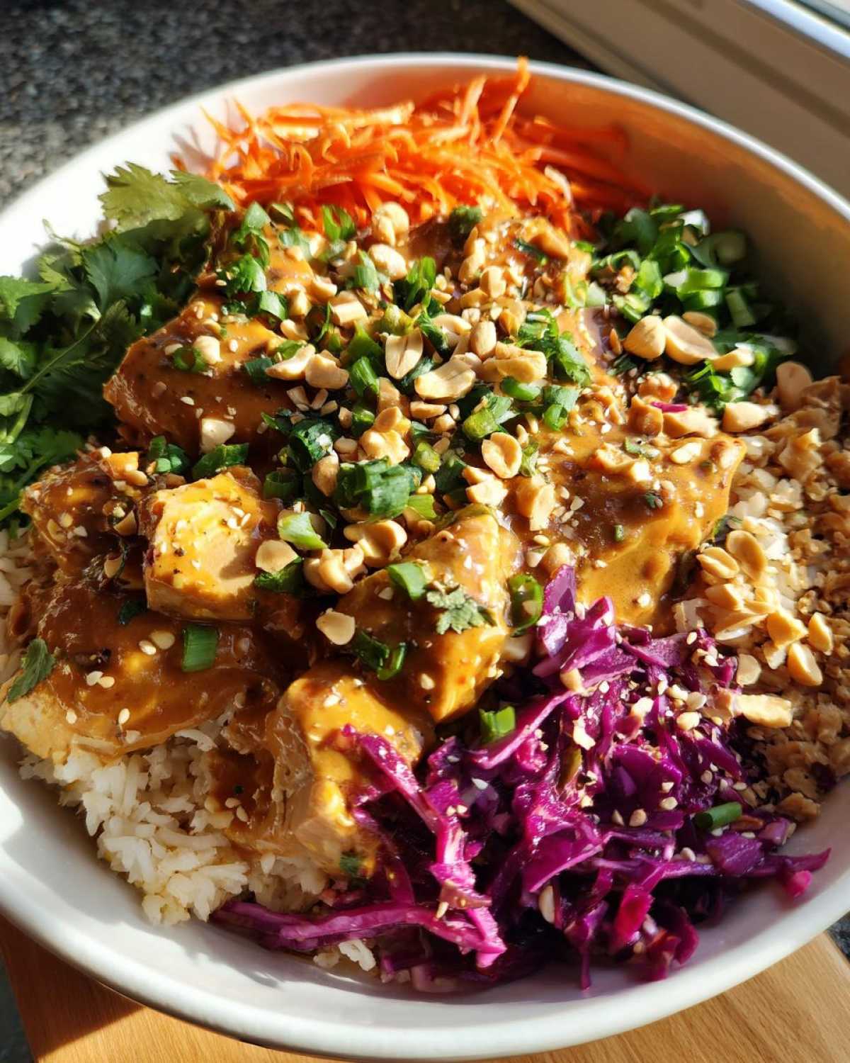 A vibrant bowl of Thai Peanut Chicken Crunch Bowls featuring saucy chicken over rice, topped with peanuts, scallions, carrots, and red cabbage.