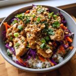 A close-up of a colorful Thai Peanut Chicken Crunch Bowls layered with rice, shredded cabbage, carrots, and topped with peanut sauce, cilantro, and chopped peanuts.