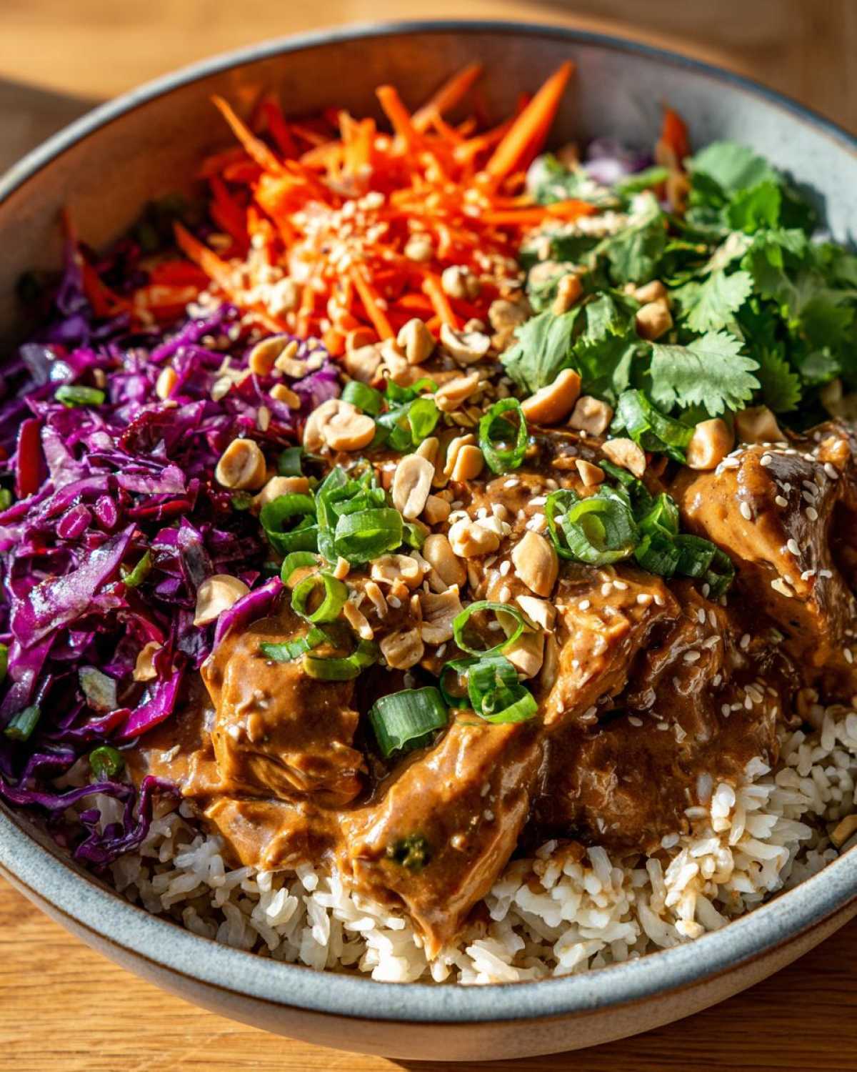 A vibrant bowl of Thai Peanut Chicken Crunch Bowls featuring peanut sauce coated chicken over rice, topped with shredded carrots, red cabbage, peanuts, and cilantro.