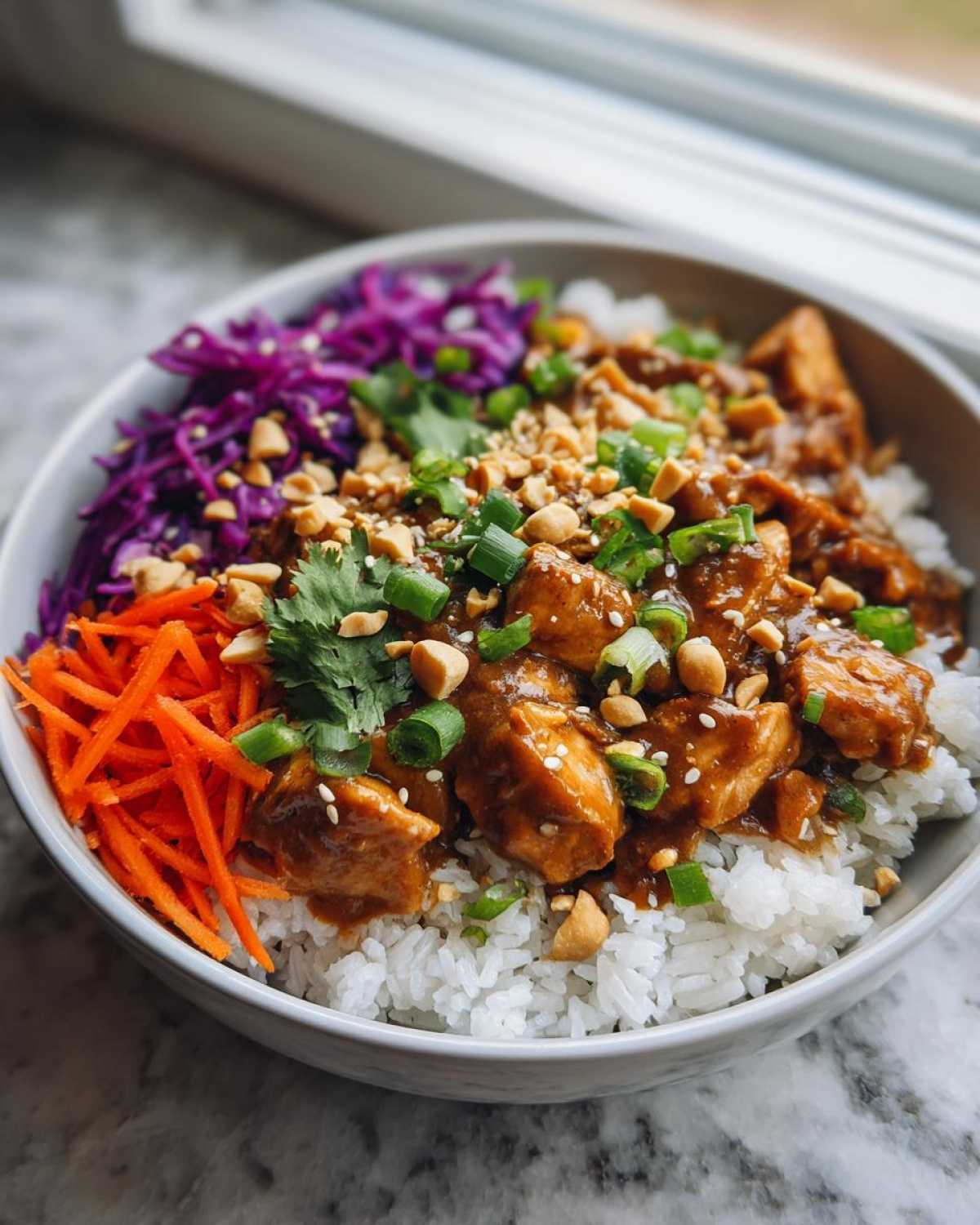 A vibrant bowl of Thai Peanut Chicken Crunch Bowls featuring saucy chicken over white rice, topped with shredded carrots, red cabbage, peanuts, and green onions.
