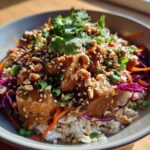 A close-up of a bowl filled with Thai Peanut Chicken Crunch Bowls, featuring saucy chicken over rice, topped with peanuts, cilantro, and slaw.