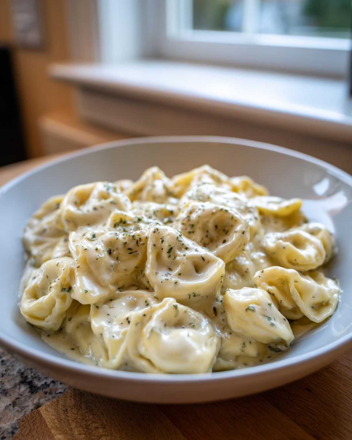 Close-up of a white bowl filled with Tortellini Alfredo Recipe covered in rich white sauce and sprinkled with herbs.
