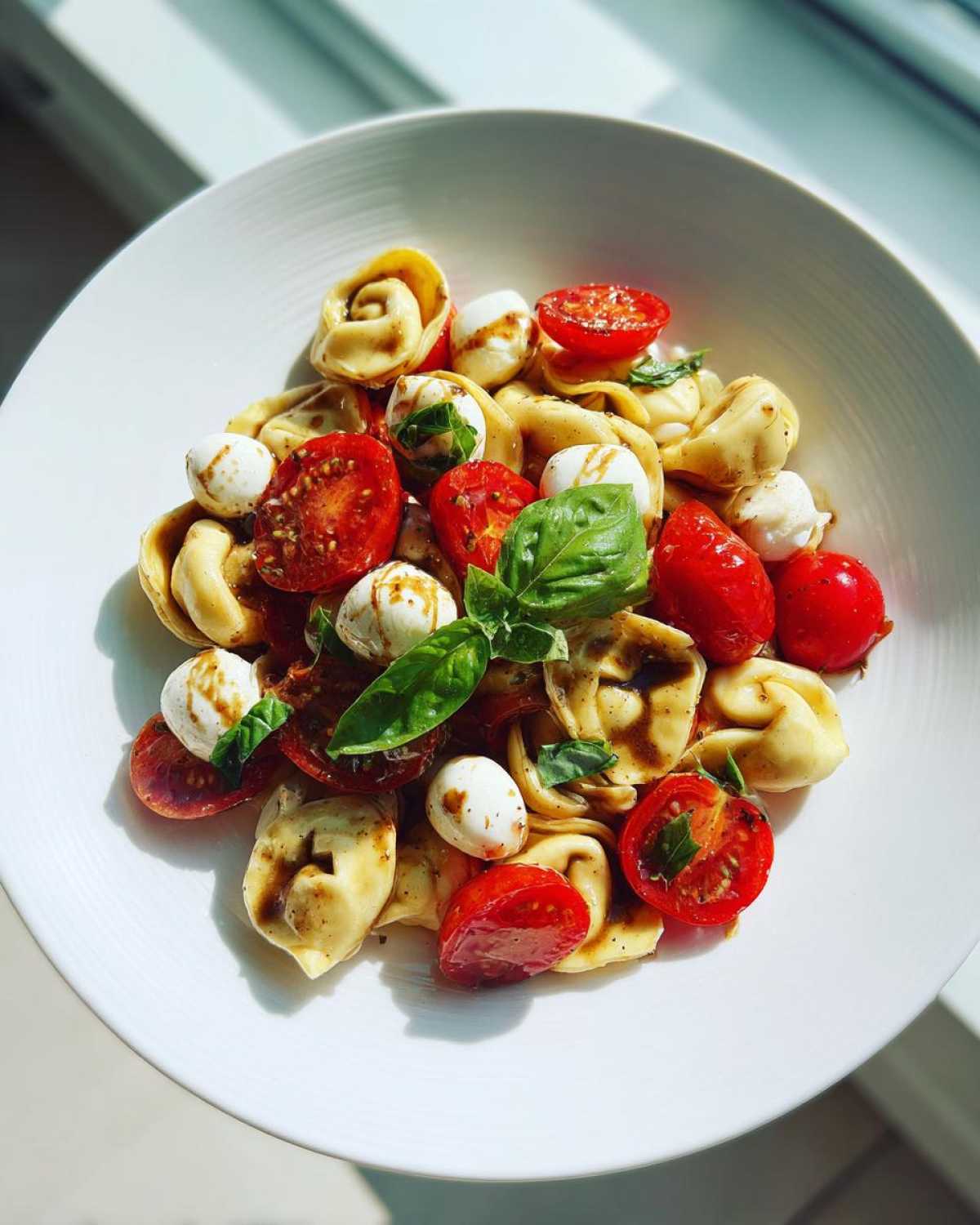 A vibrant Tortellini Caprese Salad Recipe featuring tortellini, cherry tomatoes, mozzarella balls, and basil, drizzled with balsamic glaze.