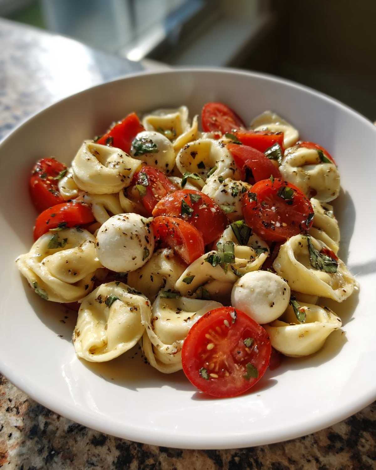 Close-up of a white bowl filled with Tortellini Caprese Salad Recipe featuring tortellini, halved cherry tomatoes, and mozzarella balls.