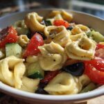 Close-up of a white bowl filled with Amazing Tortellini Pasta Salad featuring cheese tortellini, chopped tomatoes, cucumbers, and black olives.