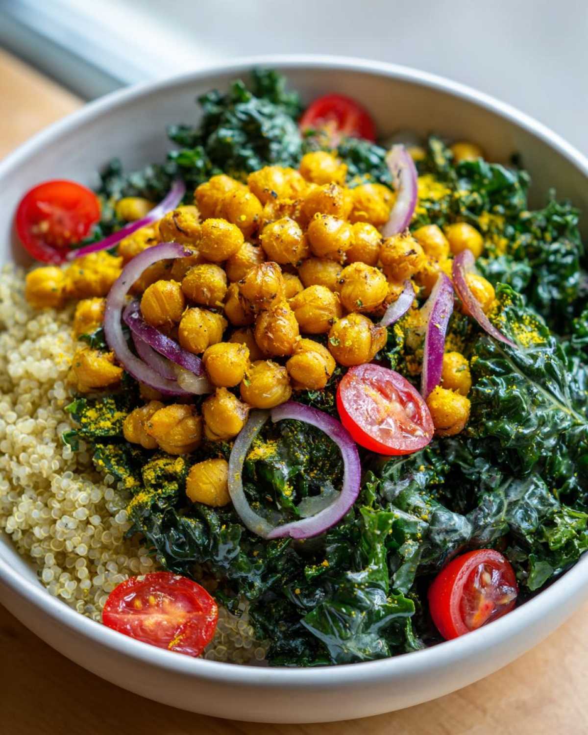 A close-up of a healthy bowl featuring quinoa, massaged kale, turmeric roasted chickpeas, red onion slices, and cherry tomatoes.