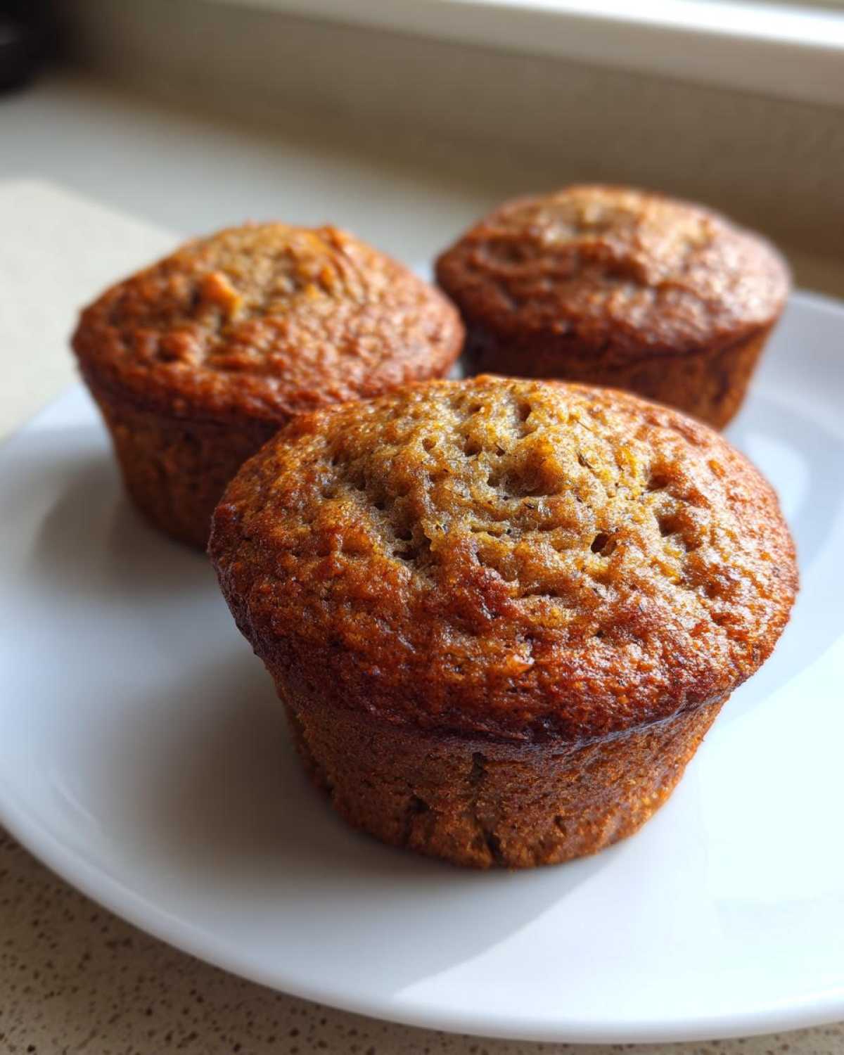 Close-up of three freshly baked Vegan banana muffins with golden brown tops resting on a white plate.