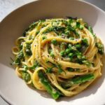 A creamy bowl of Vegan Lemon Asparagus Pasta with bright green peas, asparagus spears, and pea shoots, topped with black pepper.