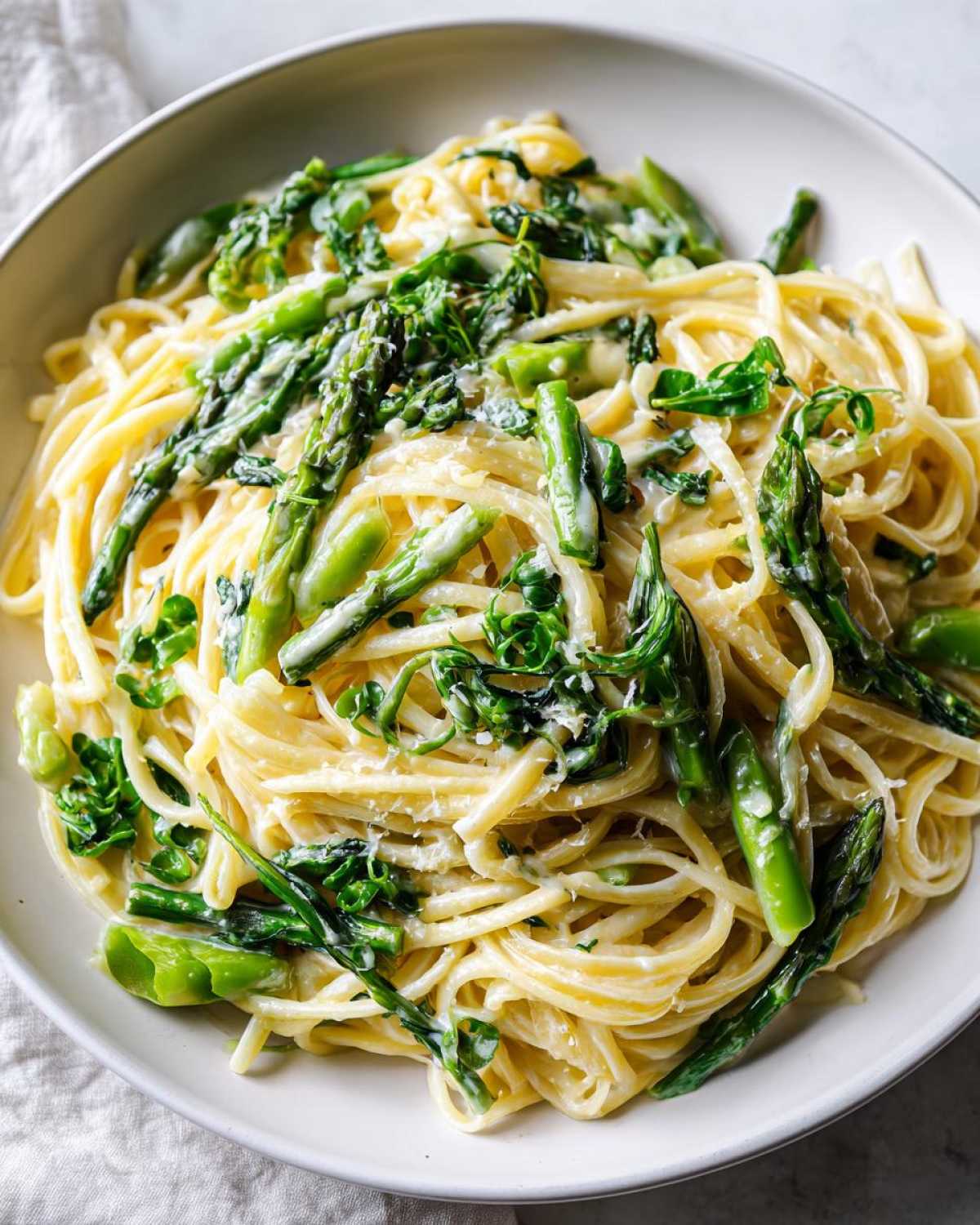 Close-up of creamy Vegan Lemon Asparagus Pasta with bright green asparagus spears and pea shoots in a white bowl.