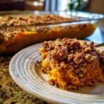 A close-up serving of bright orange Vegan squash casserole topped with a crunchy pecan crumble, with the baking dish blurred in the background.