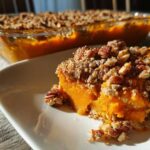 A close-up serving of bright orange Vegan squash casserole topped with a crunchy pecan crumble, with the baking dish visible in the background.