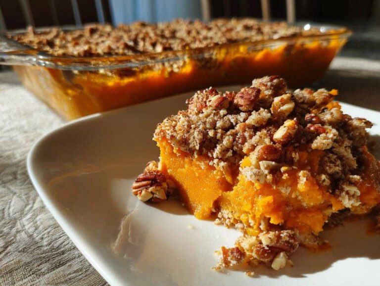 A close-up serving of bright orange Vegan squash casserole topped with a crunchy pecan crumble, with the baking dish visible in the background.