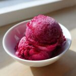 Two scoops of intensely colored, frosty Raspberry Sorbet served in a small white bowl, catching bright sunlight.