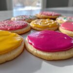 Close-up of several round sugar cookies topped with vibrant, glossy sugar cookie icing in pink, yellow, and white with sprinkles.