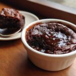 A rich, dark chocolate cobbler served warm in a white ramekin with a glossy sauce pooling on top.