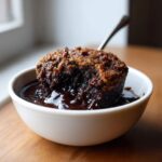A close-up of a warm chocolate cobbler with a crumbly top, sitting in a pool of rich chocolate sauce in a white bowl.