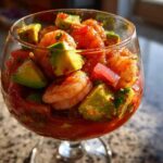 Close-up of a zesty shrimp cocktail featuring plump shrimp and chunks of avocado in a rich tomato-based sauce, served in a stemmed glass.