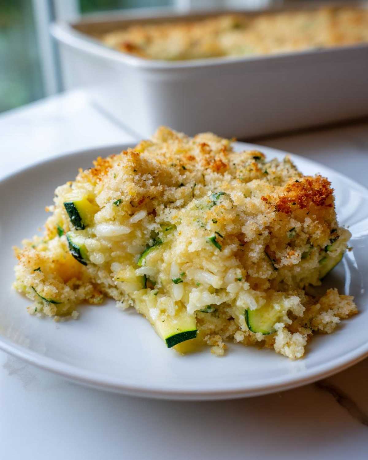 A close-up serving of Zucchini and Rice Casserole on a white plate, showing rice, zucchini pieces, and a golden, crunchy breadcrumb topping.
