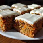 Close-up of several square Zucchini Carrot Bars recipe pieces topped with thick white cream cheese frosting on a white plate.