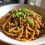 A close-up shot of glossy, thick Asian Garlic Noodles coated in a dark sauce and topped with fresh green onions.