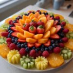 A vibrant, circular Baby Bloom Fruit Platter featuring cantaloupe, strawberries, blueberries, raspberries, oranges, and carved kiwi and pineapple flowers.