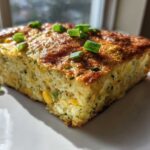 A golden-brown square slice of Baked Zucchini and Corn Casserole topped with fresh green onions on a white plate.