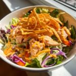 A vibrant bowl of Bang Bang Salmon Salad featuring flaked salmon, mixed greens, red cabbage, and crispy wonton strips drizzled with orange sauce.
