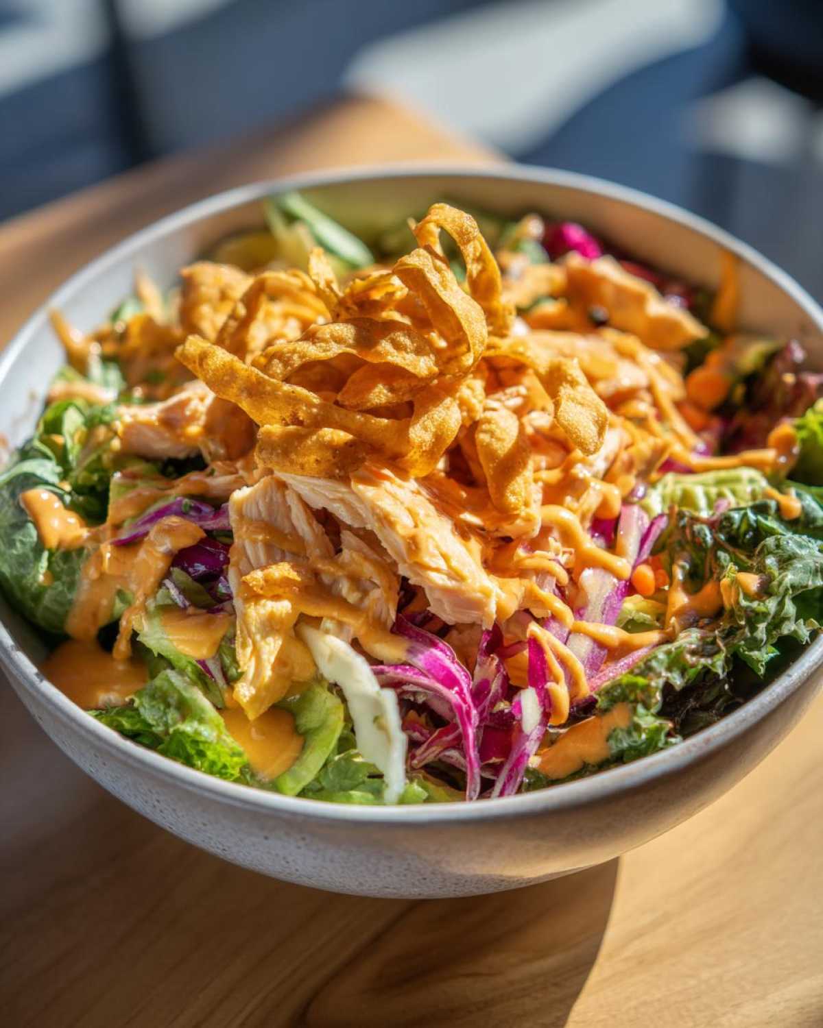 A close-up bowl of Bang Bang Salmon Salad featuring flaked salmon, mixed greens, purple cabbage, and drizzled with orange sauce, topped with crispy wonton strips.