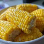 Close-up shot of several pieces of bright yellow Buttered Corn on the cob piled in a white bowl.