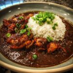 A close-up of rich, dark Chicken Étouffée served with a mound of white rice and garnished with chopped green onions.