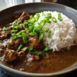Close-up of a bowl of rich, dark Chicken Étouffée served next to a mound of white rice and topped with fresh green onions.