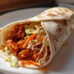 Close-up of a perfectly rolled Chili Chicken Wraps Lunch filled with saucy chicken pieces and crisp slaw on a white plate.