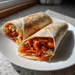 Two tightly rolled Chili Chicken Wraps Lunch halves filled with spicy chicken and shredded carrots/cabbage on a white plate.
