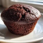 Close-up of a dark, moist chocolate muffin served on a white plate, perfect pairing for Recetas de Café.