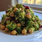 A vibrant mound of Chopped Broccoli and Chickpea Salad featuring broccoli florets, chickpeas, dried cranberries, and seeds on a white plate.
