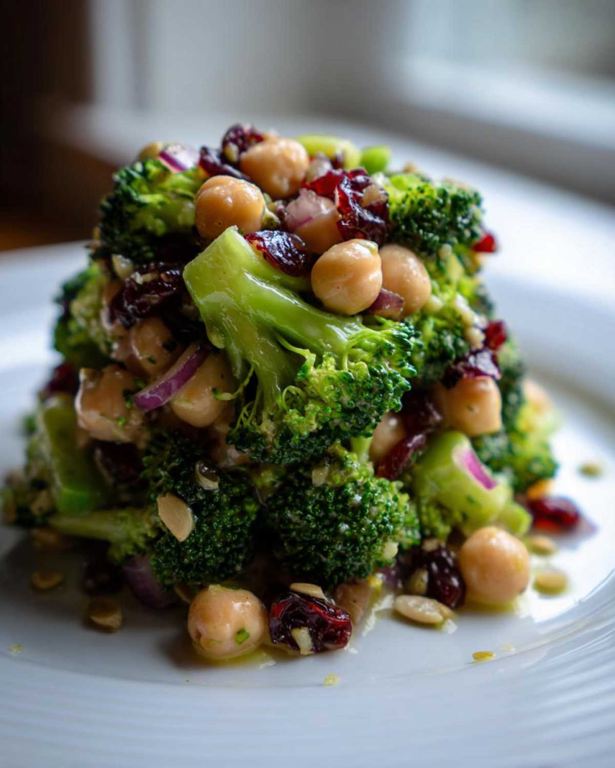 A vibrant mound of Chopped Broccoli and Chickpea Salad featuring bright green florets, chickpeas, dried cranberries, and red onion on a white plate.